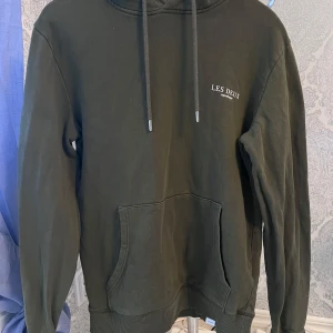 Grön hoodie från Les Deux - Säljer en stilren grön hoodie från Les Deux med justerbar dragsko i huvan och en praktisk magficka. Perfekt för en avslappnad look. Märkeslogga broderad på bröstet.