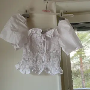 Säljer en söt vit blus från NA-KD med puffiga korta ärmar och smockdetaljer. Blusen har en knytning framtill och volangkant nedtill. Perfekt för en somrig look! Använd max 1 gång 