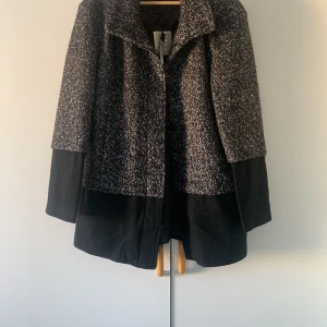 Svart och vit kappa från VILA - Säljer en elegant svart och vit kappa från VILA. Den har en stilren design med en mix av svart och salt & peppar-mönster. Perfekt för att hålla stilen under kyligare dagar.