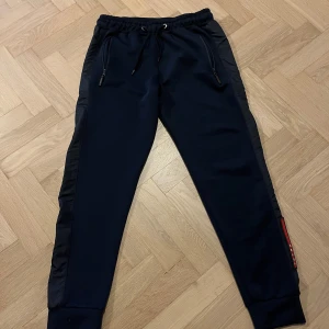 Prada byxor - Snygga mörkblå mjukisbyxor från Prada med elastisk midja och dragsko. Byxorna har svarta sidopaneler och en diskret logga. Perfekta för en avslappnad stil med en touch av lyx.