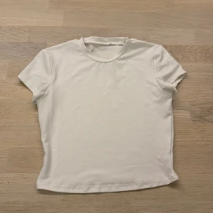 Vit t-shirt i polyester - Säljer en enkel och stilren vit t-shirt i polyester med korta ärmar. Perfekt för en minimalistisk look. T-shirten har en rund halsringning och är tillverkad i ett mjukt och stretchigt material. Rött så cropped och ganska tajt i formen. Är osäker vart den är ifrån eftersom att den saknar lappar. 