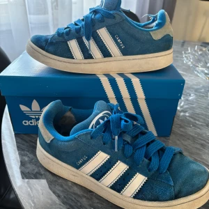 Blå Adidas Campus sneakers - Snygga blå Adidas Campus sneakers med vita detaljer och klassiska tre ränder på sidan. Skorna har en bekväm passform och är perfekta för en avslappnad stil. De är tillverkade i mocka och har snörning framtill.
