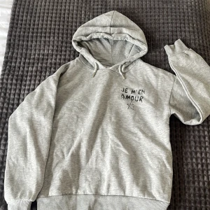 Grå hoodie från Gina Tricot - Säljer en grå hoodie från Gina Tricot med texten 'Je m'en amour'. Hoodien har en klassisk design med huva och ribbade muddar vid ärmslut och nederkant. Använda få gånger och inga fläckar!!❣️❣️❣️