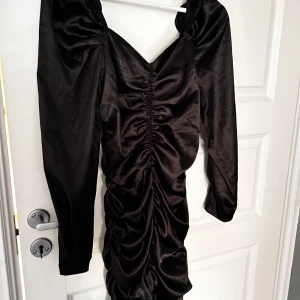 Svart långärmad klänning - Elegant svart klänning med lång ärm och rynkade detaljer framtill. Klänningen har en v-ringad design och är tillverkad i ett glansigt material som ger en lyxig känsla.