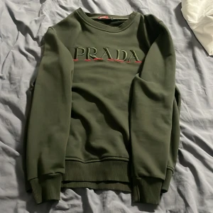 Grön tröja från Prada ifrån farfetch - Säljer en stilren grön tröja från Prada med broderad logotyp på framsidan. Tröjan har långa ärmar och ribbade muddar vid ärmslut och nederkant. Perfekt för en avslappnad men ändå lyxig look. 