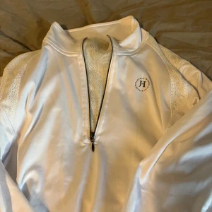 Vit tröja med dragkedja från JH Collection - Säljer en stilren vit tröja från JH Collection med dragkedja framtill. Perfekt att ha till hopp eller dressyr tävling. Den har ett skönt flis material inuti och har inga tecken på att den har används en gång.