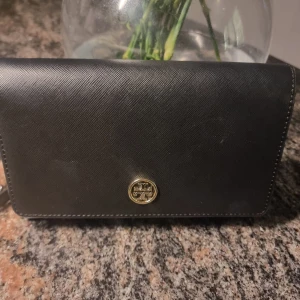 Svart väska från Tory Burch - Elegant svart väska från Tory Burch i skinn med en stilren design. Plånboken har en guldlogga på framsidan och flera kortfack inuti. Perfekt för att hålla ordning på dina kort och kontanter.