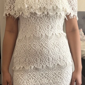 Vit spetsklänning - Säljer en elegant vit spetsklänning med offshoulder-design och vackra spetsdetaljer. Klänningen har en kort längd och en figurnära passform som framhäver silhuetten. Perfekt för speciella tillfällen!