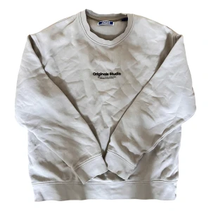Beige sweatshirt från Jack & Jones - Säljer en stilren beige sweatshirt från Jack & Jones med texten 'Originals Studio' tryckt på bröstet. Tröjan har en klassisk rund hals och långa ärmar. Perfekt för en avslappnad look.