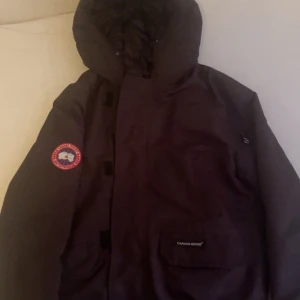 Svart parkajacka från Canada Goose - Säljer en svart parkajacka från Canada Goose med deras ikoniska märke på ärmen. Jackan har en långärmad design och är perfekt för kalla säsonger. Den är tillverkad i slitstarkt material och har en praktisk huva för extra skydd mot väder och vind. Lite trasigt drag kedja (metallen) men går ändå att dra upp helt 😁
