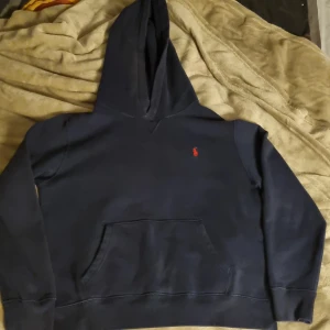 Mörkblå hoodie från Ralph Lauren - Säljer en stilren mörkblå hoodie från Ralph Lauren med det klassiska röda logotypen på bröstet. Tröjan har en bekväm passform med långa ärmar och en praktisk magficka. Perfekt för en avslappnad look. Storlek L i barnstorlek xs i vuxen.