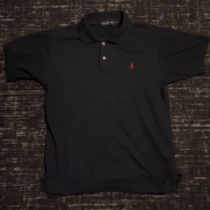 Pikétröja från Ralph Lauren - Säljer en klassisk pikétröja från Ralph Lauren med röd broderad logga på bröstet. Tröjan har korta ärmar och knappar vid kragen. Perfekt för en stilren look.