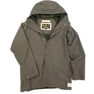 Regn jacka Barbour. - Säljer en stilren grå regnjacka från Barbour med huva och knappar. Jackan har en klassisk design med praktiska fickor och är perfekt för regniga dagar. Den är tillverkad i ett slitstarkt material som håller dig torr och bekväm.