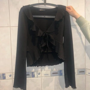 Svart bolerotopp från Gina Tricot - Snygg svart bolerotopp från Gina Tricot med volangdetaljer och knyt framtill. Toppen har långa ärmar och en croppad passform som ger en trendig look. Perfekt för att lägga till en touch av stil till din outfit.