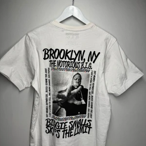 Vit t-shirt med The Notorious B.I.G. tryck - Säljer en vit t-shirt med tryck av The Notorious B.I.G. och texten 'Brooklyn, NY' samt 'Biggie Smalls Sky's the Limit'. Perfekt för fans av hip-hop och streetwear.