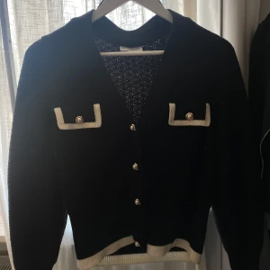 Svart kofta från Holly & Whyte - Snygg svart kofta från Holly & Whyte , aldrig använd nyligen köpt för 500kr. Med vita detaljer och pärlknappar. Koftan har två dekorativa fickor framtill och ribbade kanter. Perfekt för en stilren look.