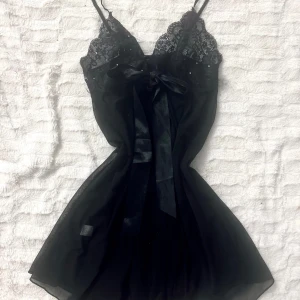 Svart spetsklänning med rosett - Elegant svart klänning med spetsdetaljer och en söt rosett framtill. Axelbandslös design med en luftig och lätt känsla. Perfekt för en stilfull kväll. Obs under rosetten (precis under) finns ett knyte som har gått upp lite på bandet. Går att gömma under rosetten. 