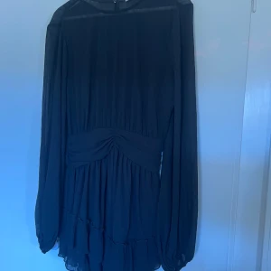 Svart klänning med volangdetaljer - Elegant svart klänning med långa ärmar och volangdetaljer i nederkant. Klänningen har en åtsittande midja med rynkade detaljer som ger en smickrande siluett. Perfekt för en stilren look.