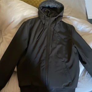 Svart jacka från Michael Kors - Snygg svart jacka från Michael Kors med huva och dragkedja. Jackan har en mjuk insida och stora bokstäver på ärmen. Väldigt bekväm och lätt att matcha till sin outfit. 