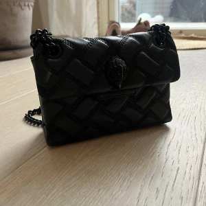 Svart quiltad axelväska från Kurt Geiger - Snygg svart axelväska från Kurt Geiger med quiltad design och kedjerem. Väskan har en dekorativ detalj på framsidan och är perfekt för att ge en stilren touch till din outfit.