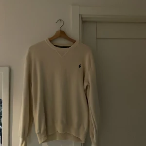 Beige tröja från Ralph Lauren - Säljer en stilren beige tröja från Ralph Lauren med rund hals och broderad logotyp på bröstet. Tröjan har långa ärmar och ribbade muddar vid ärmslut och nederkant. Perfekt för en avslappnad och klassisk look. Den har blivit för liten.