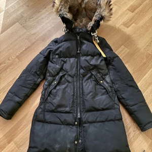 Svart dunjacka med pälsfodrad huva - Säljer en svart dunjacka från Parajumpers med en lyxig pälsfodrad huva. Jackan har en dragkedja och knappar framtill samt flera praktiska fickor. Perfekt för kalla vinterdagar. Ett snöre som man spänner till jackan där nere har gått sönder lite. 