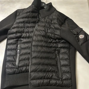  Moncler  cardigan - Säljer en stilren svart pufferjacka från Moncler med dragkedja och ribbade detaljer. Jackan har en broderad logotyp på ärmen och praktiska fickor med dragkedjor. Perfekt för kyligare dagar! den är L o m i storlek