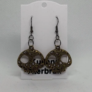 Bronsfärgade dödskalleörhängen - Unika bronsfärgade örhängen med dödskallemotiv. De har en detaljrik design med utskurna mönster och hängs på krokar. Perfekta för att ge en edgy touch till din stil.