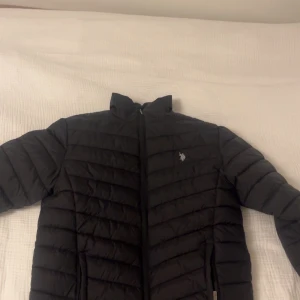 Svart pufferjacka från U.S. Polo Assn. - Säljer en svart pufferjacka från U.S. Polo Assn. med broderad logga på bröstet. Jackan har en dragkedja framtill och är perfekt för kyligare dagar. Den är stilren och enkel att matcha med olika outfits.
