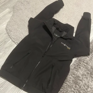 Svart Gore-Tex jacka från Sail Racing - Säljer en svart Gore-Tex jacka från Sail Racing. Jackan har dragkedja framtill och två praktiska fickor med dragkedjor. Den är designad för att stå emot väder och vind, perfekt för utomhusaktiviteter. Logotypen är broderad på bröstet och ryggen. Inte synligt använd