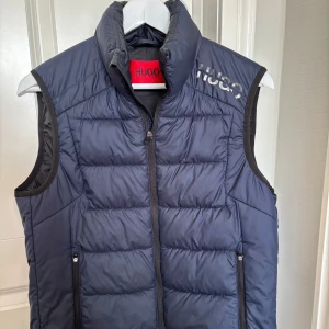 Blå dunväst från Hugo Boss - Snygg blå dunväst från Hugo Boss med dragkedja och hög krage. Västen har ett stilrent tryck på bröstet och praktiska fickor med dragkedja. Perfekt för lager-på-lager under kyliga dagar.