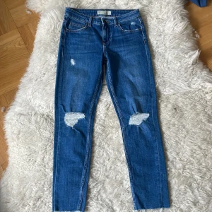 Blå jeans från Topshop st.26 - Snygga blå jeans från Topshop med slitna detaljer på knäna. De har en klassisk femficksdesign och knappgylf. Perfekta för en avslappnad stil.