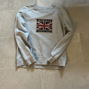 Grå sweatshirt från Morris - Säljer en stilren grå sweatshirt från Morris med ett tryck av den brittiska flaggan och texten 'Morris Since 1968' på bröstet. Tröjan har långa ärmar och en rund halsringning. Perfekt för en avslappnad stil.