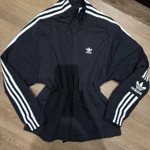 Adidas jacka - Snygg svart vindjacka från Adidas med de klassiska tre vita ränderna längs ärmarna. Jackan har dragkedja framtill och Adidas-loggan på bröstet och ärmen. Perfekt för en sportig look! Använd 2-3 ggr. Inga  detekter, skador etc 