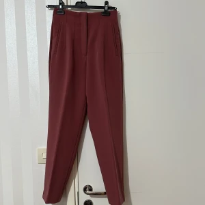  kostymbyxor med hög midja - Säljer ett par stilrena kostymbyxor i en dämpad rosa nyans. Byxorna har hög midja, raka ben och diskreta fickor framtill. Perfekta för dig som vill ha en dressad look med modern touch.