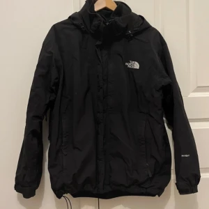 Svart vindjacka från The North Face - Svart vindjacka från The North Face med huva och dragkedja framtill. Jackan har logga på bröstet och ryggen samt flera fickor. Perfekt för blåsiga dagar och enkel att matcha med olika outfits. Fläck på armbågen