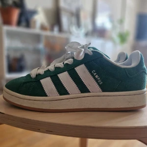 Adidas Campus gröna sneakers - Säljer ett par gröna Adidas Campus sneakers med vita ränder och vit sula. Skorna har klassisk snörning och är tillverkade i mocka med Adidas-logga på plösen. Perfekta för dig som gillar retrostil och streetwear.