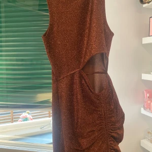 Brun glittrig ärmlös klänning med cut out - Säljer en brun glittrig klänning med ärmlös design och snygg cut out-detalj i midjan. Klänningen har åtsittande passform och lätt rynkade partier på sidan. Perfekt för dig som vill sticka ut!