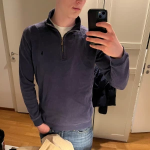 Mörkblå tröja med dragkedja från Polo Ralph Lauren - Säljer en mörkblå långärmad tröja från Polo Ralph Lauren med halv dragkedja och liten broderad logga på bröstet. Tröjan har hög krage och är perfekt för en stilren och avslappnad look.