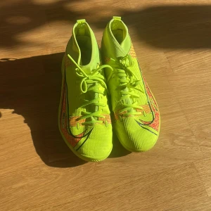 Neongröna fotbollsskor från Nike - Snygga neongröna fotbollsskor från Nike med svart och röd grafik. Skorna har snörning och en hög ankelkrage för extra stöd. Perfekta för att dominera på planen med stil.
