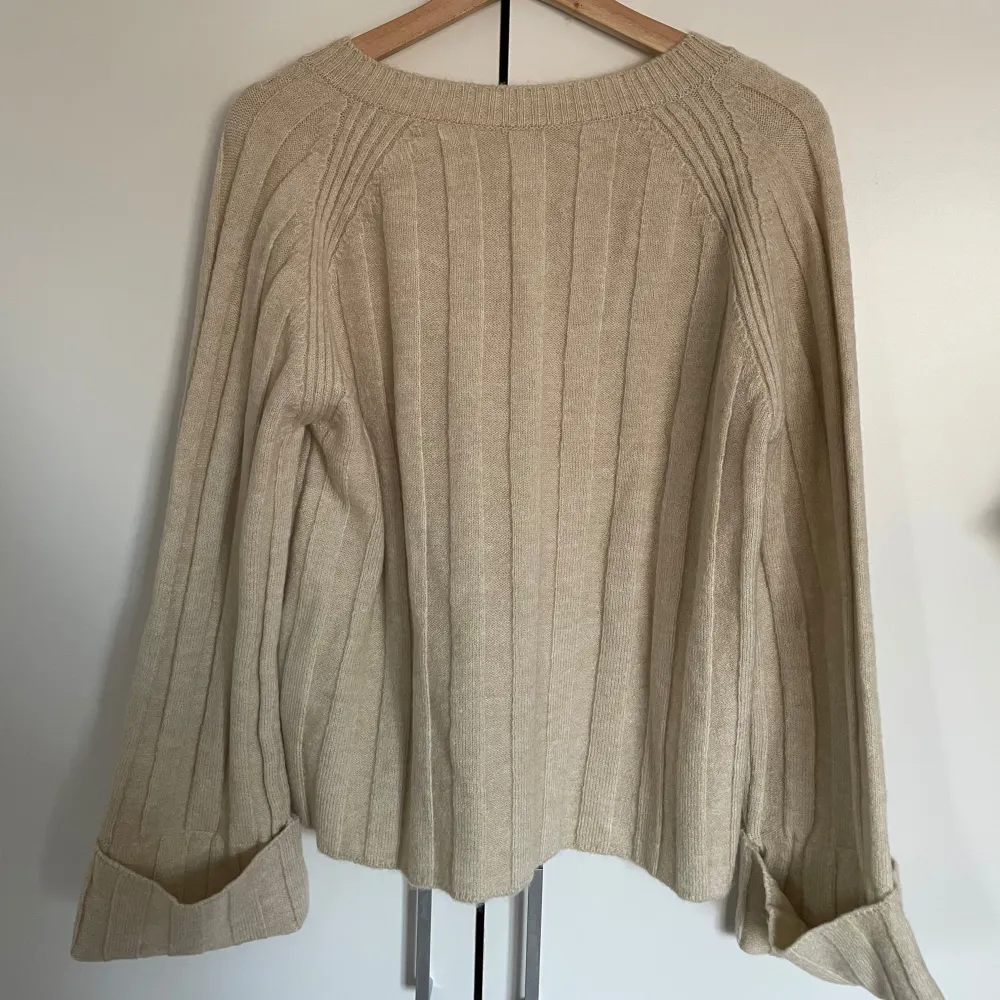 Mysig beige stickad tröja med v-ringad hals och ribbat mönster. Tröjan har långa ärmar med uppvikta muddar och en avslappnad passform.. Neuletakit & Villapaidat.