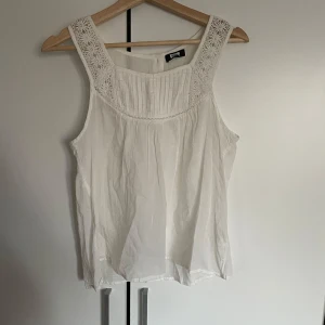 Vit ärmlös blus med spetsdetaljer från Bik Bok - Säljer en söt vit ärmlös blus från Bik Bok i storlek S. Blusen har breda axelband med spetsdetaljer och knappar i ryggen.