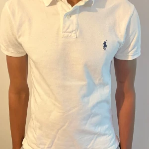 Vit piké från Polo Ralph Lauren - Klassisk vit pikétröja från Polo Ralph Lauren med blå broderad logga på bröstet. Slim fit-modell med korta ärmar och krage med knappar. Perfekt för en clean och stilren look. Den är köpt på zalando för 1200kr. Vid frågor eller funderingar är det bara att höra av sig.