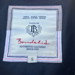 Mörkblå kavaj från Bondelid - Säljer en stilren mörkblå kavaj från Bondelid med två knappar framtill och broderad logga på bröstet. Kavajen har lång ärm och en patch med amerikansk flagga på ena ärmen. Perfekt för dig som gillar klassisk stil.