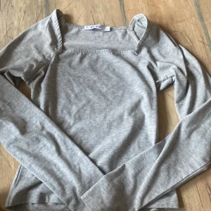 Grå långärmad topp från NA-KD - Säljer en stilren grå långärmad topp från NA-KD. Toppen har en fyrkantig ringning och är tillverkad i mjukt material som sitter snyggt på kroppen. Perfekt att matcha med jeans eller kjol!