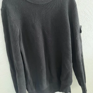 Svart stickad tröja från Stone Island - Snygg svart stickad tröja från Stone Island med deras ikoniska märke på ärmen. Perfekt för kyligare dagar med sin långärmade design och klassiska look.