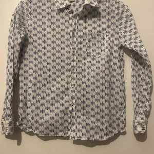 Vit skjorta med blått mönster från Massimo Dutti - Säljer en vit skjorta från Massimo Dutti med blått grafiskt mönster. Skjortan har lång ärm, klassisk krage och knäppning framtill. Perfekt för dig som gillar stilrena men ändå unika plagg!