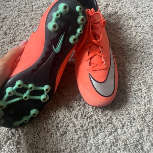 Nike fotbollsskor - Säljer ett par orangea Nike Mercurial fotbollsskor med svart och silverdetaljer. Skorna har snörning och är designade för spel på gräs med dobbar undertill. Perfekta för dig som vill sticka ut på planen!