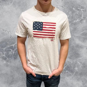 Ralph Lauren T-shirt vit USA Flag - •Ralph Lauren T-shirt USA Flag🇺🇸 vit. Skick: 7-8/10, Inga skador! slitningarna i flaggan är original design! Lite stor i halsen men annars inga konstigheter. Storlek: 14/16 yrs= strl S~. Modell: 173 cm, 65 kg. Sitter bra på modellen på bild! Authentic: ✅. Hör av dig vid frågor eller funderingar. Mvh Hessler’s;) 