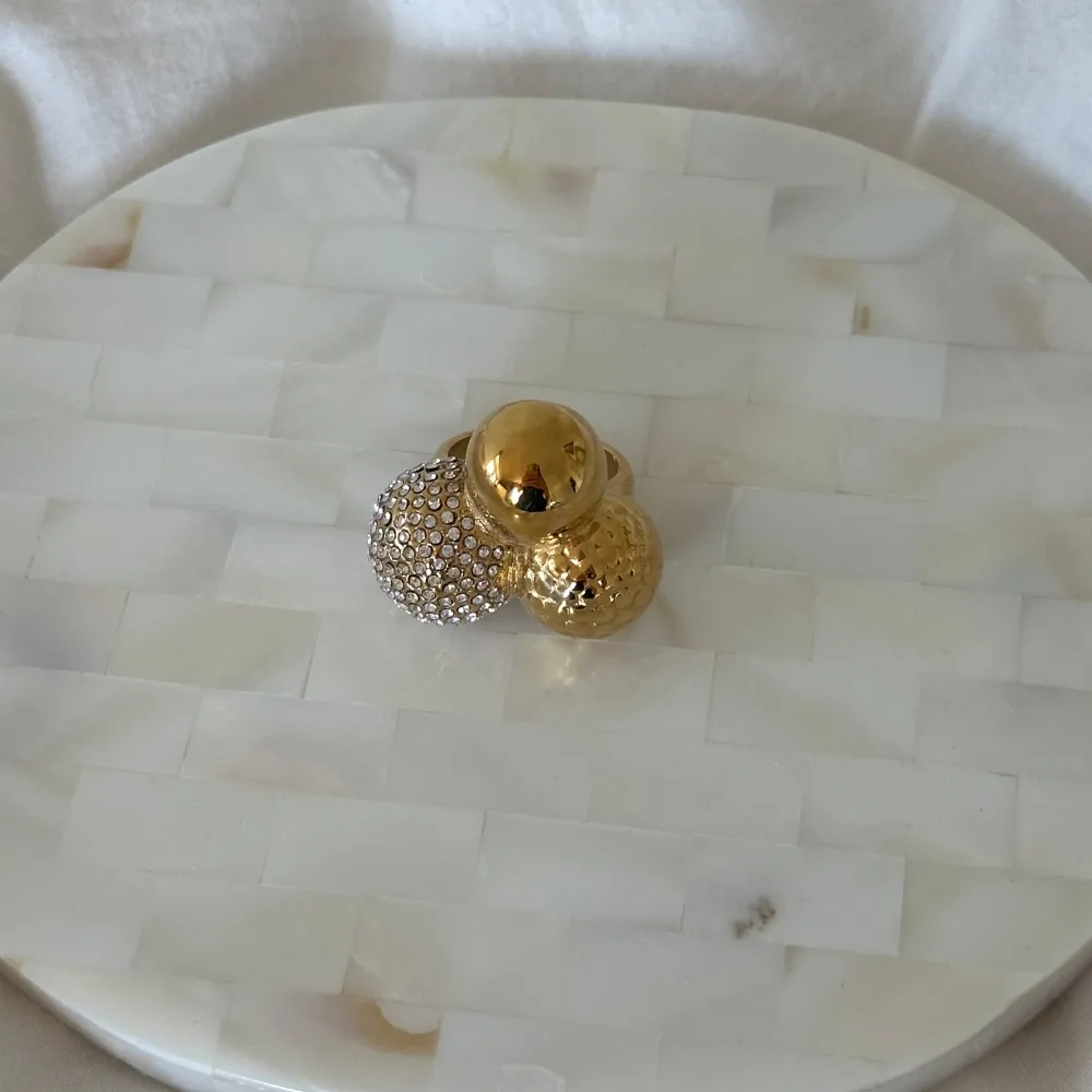 Snygg guldfärgad ring med tre stora kulor, varav en är täckt med glittrande stenar. Perfekt för att ge en extra touch av glamour till din outfit. Guld plågering med stainless steel 🤍. Asusteet.